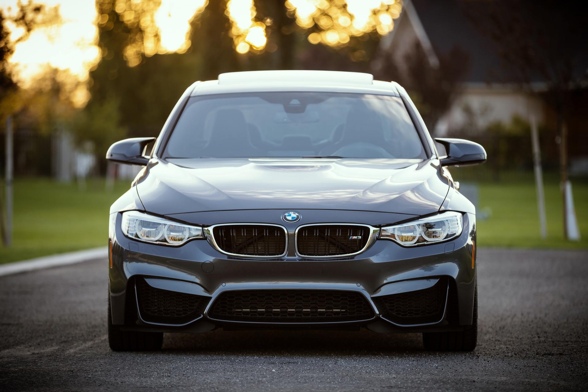 Dark gray BMW M3 on a paved road, facing forward. Sunset in the background.