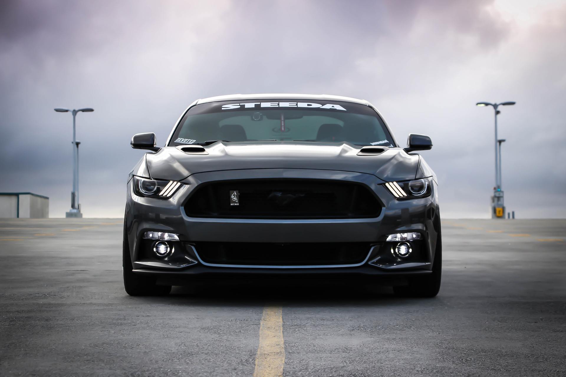 Black Ford Mustang, front view, on a rooftop parking lot under a cloudy sky.