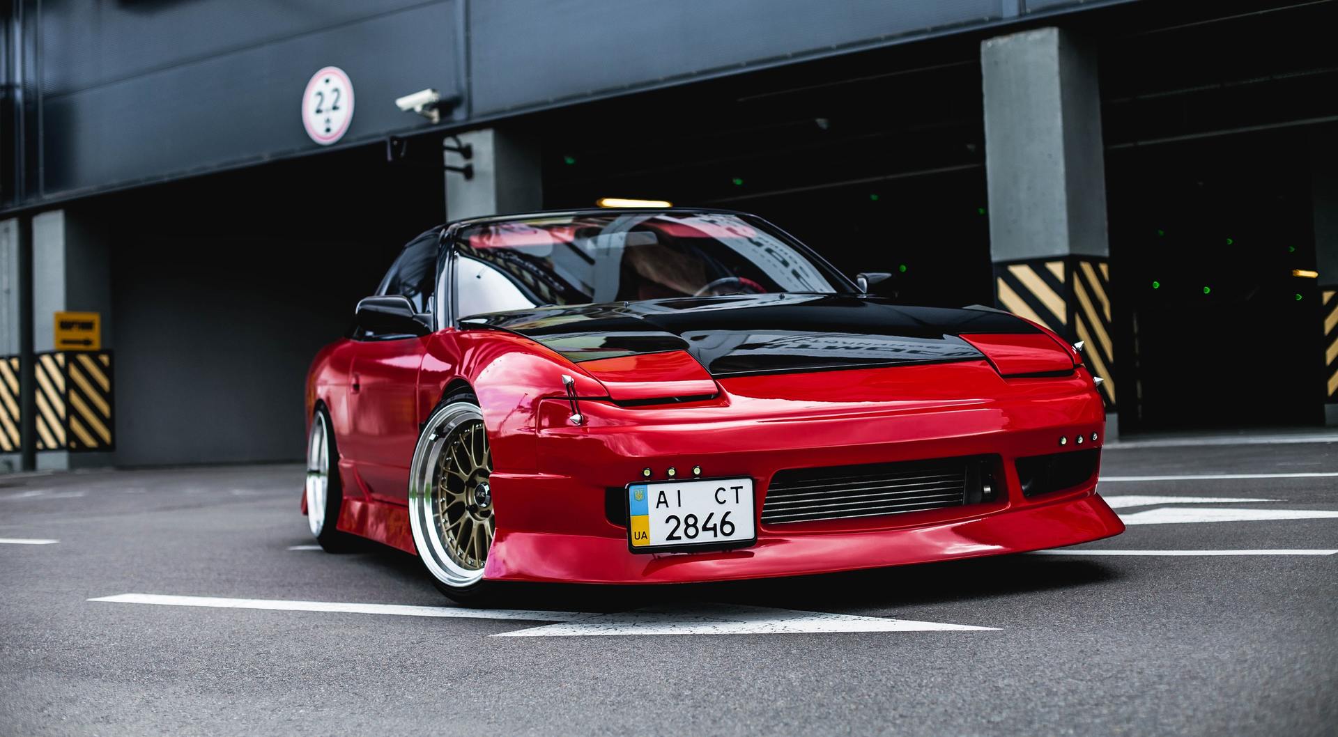 Red Nissan 240SX modified with a black hood, parked in a parking garage.