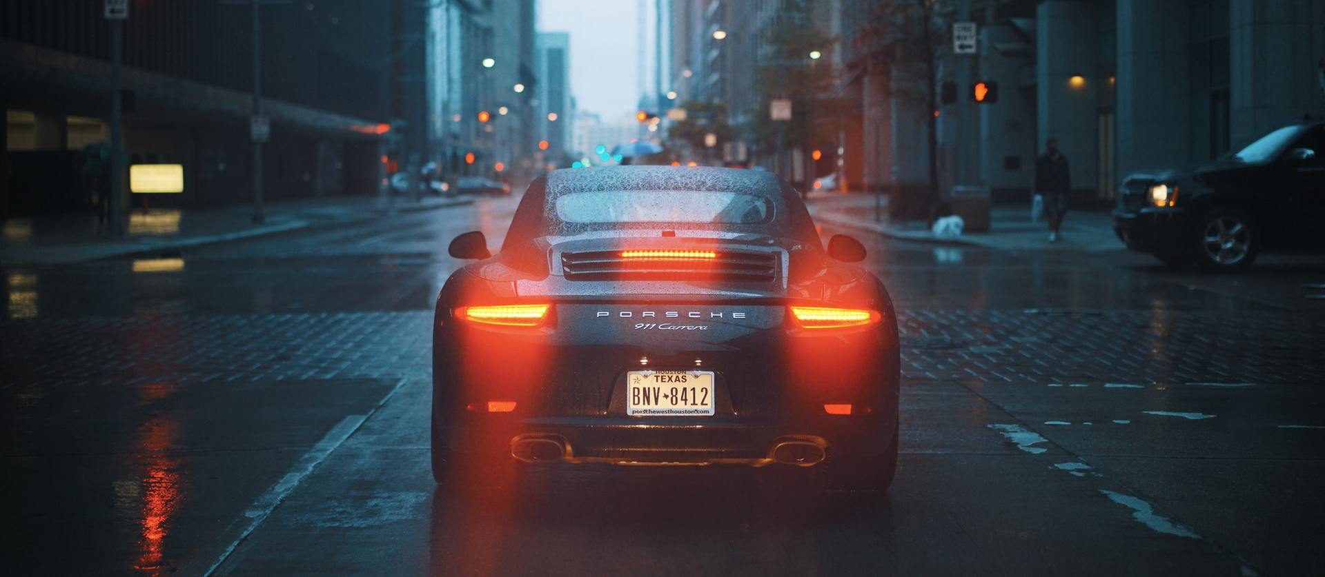 Black sports car driving in the rain down a city street, rear lights glowing red, and wet pavement reflecting light.